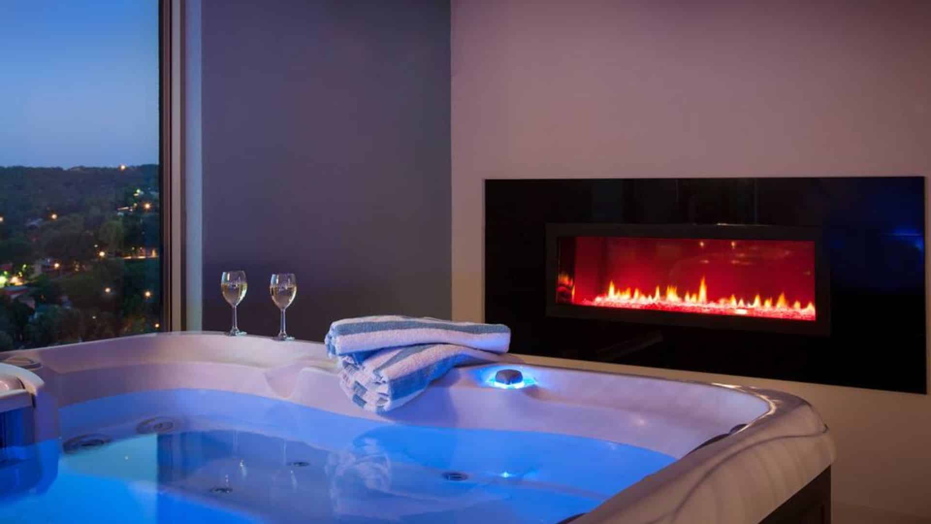 A luxurious hot tub with glowing blue water, two wine glasses, and a modern fireplace in a stylish room.