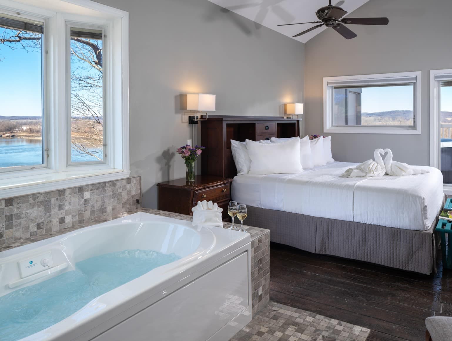 A cozy hotel room featuring a bed, a Jacuzzi, and windows with a view of a lake.