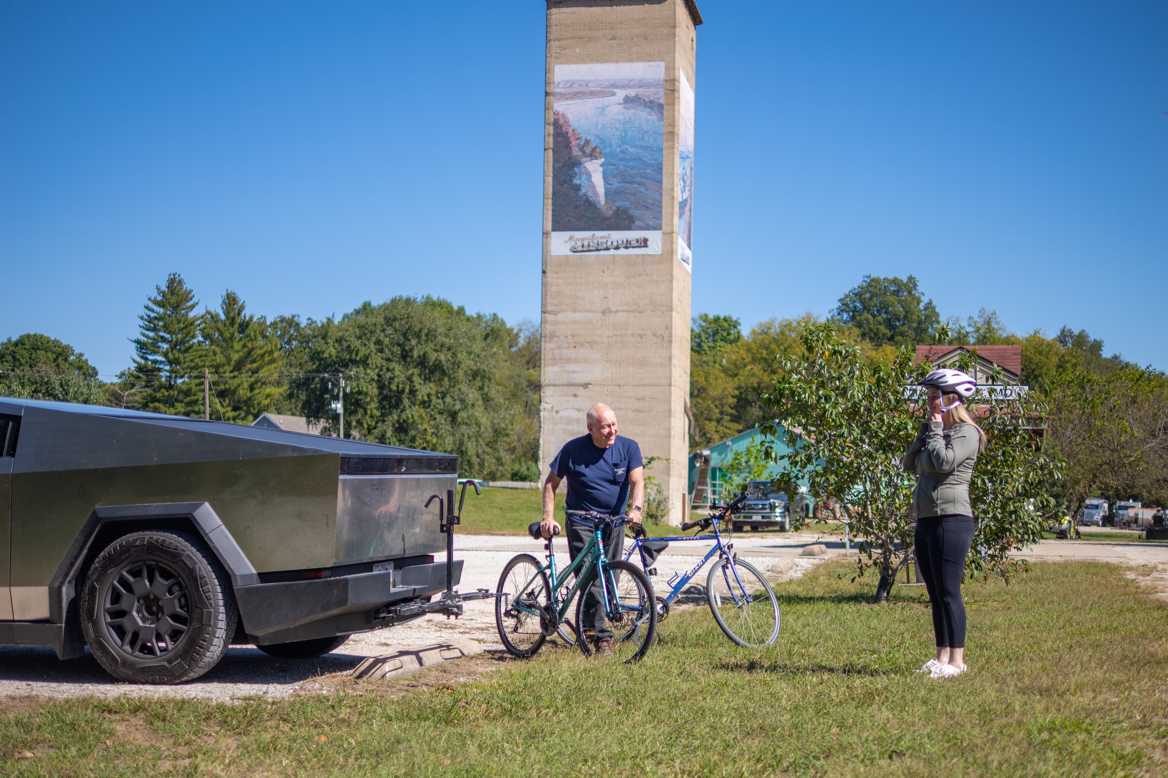 On the edge of a gravel parking lot a man is wheeling a bicycle over to a woman standing in the grass putting her helmet on. There is a second bicycle in the grass. The man is stepping away from a Cybertruck.