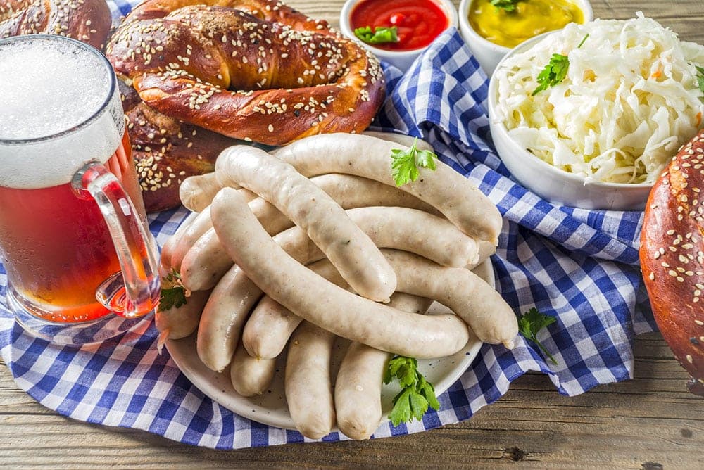A plate of white sausages with pretzels, sauerkraut, and a glass of beer on a checkered cloth.