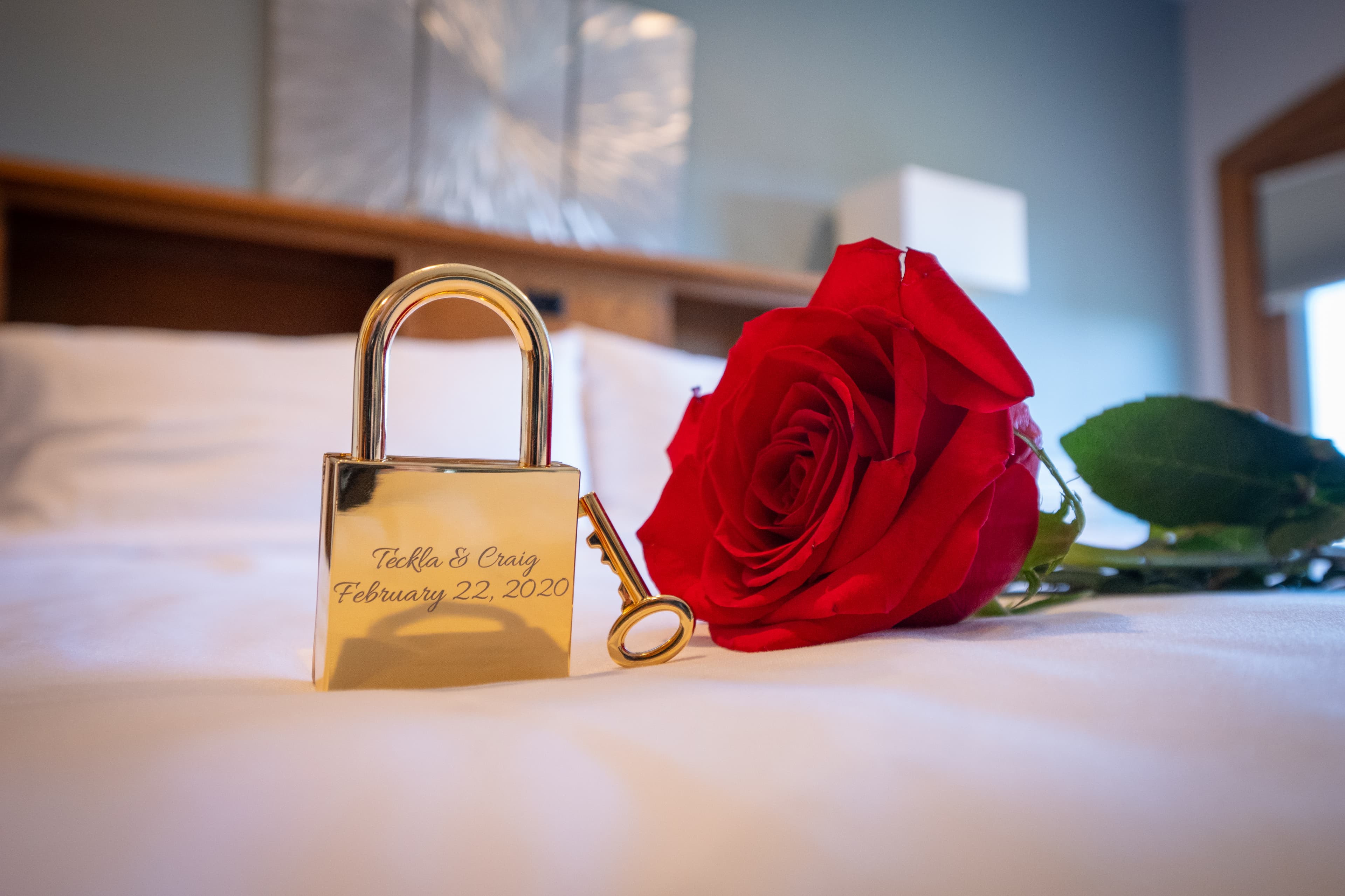 A close up photo of a gold padlock, key and red rose are on a bed with white linens.
