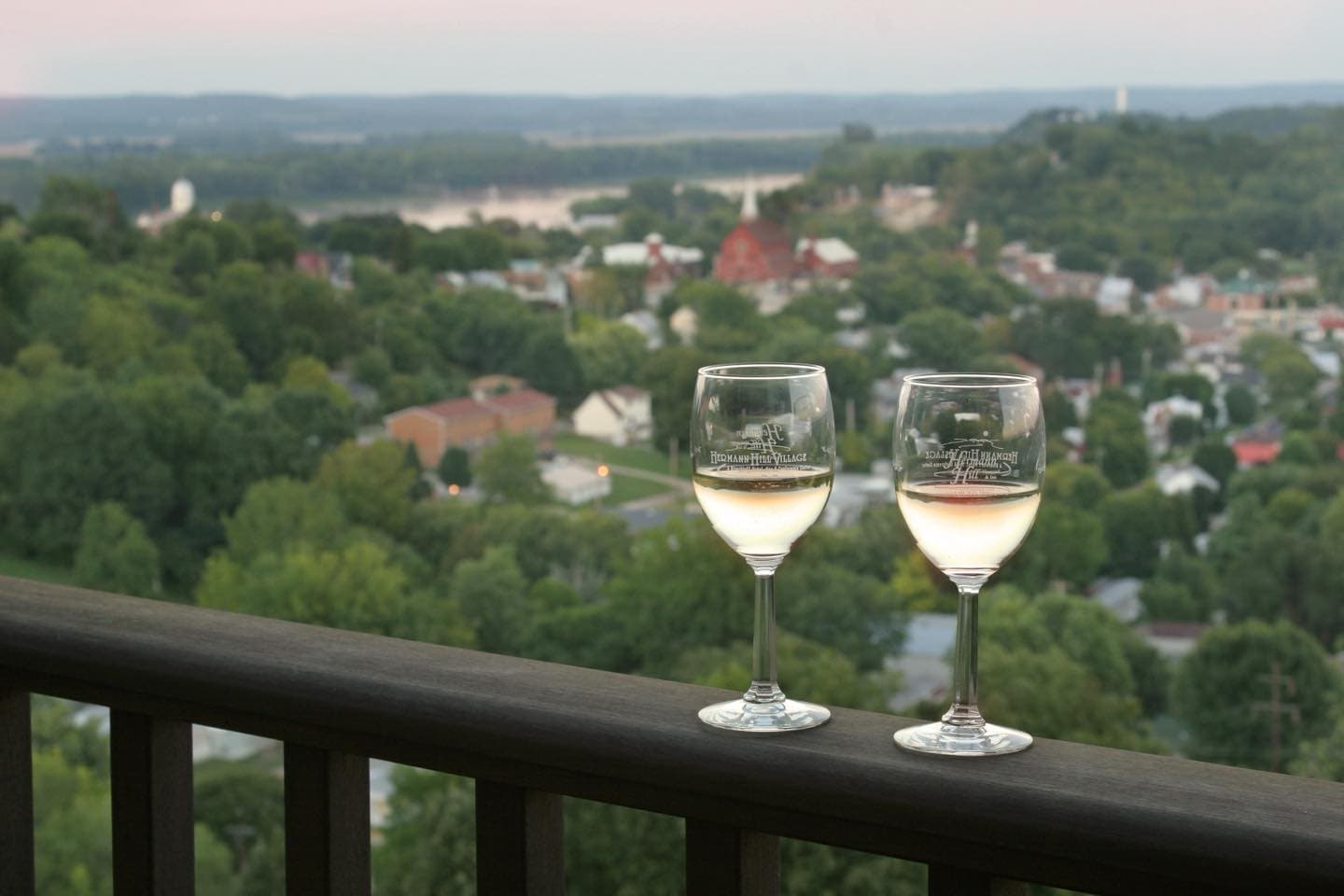 Two glasses of white wine on a balcony overlooking a lush green valley and small town.
