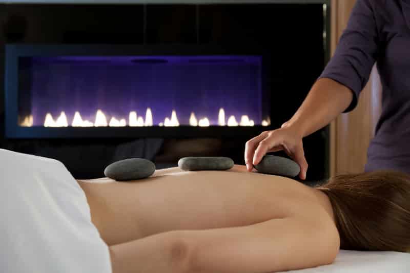 A person receives a hot stone massage with three stones placed on their back in front of a fireplace.