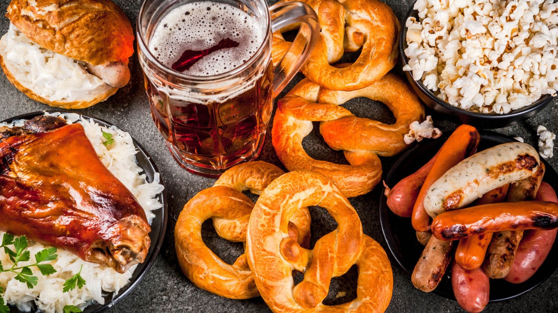 A variety of snacks including pretzels, popcorn, sausages, and a beer in a glass.