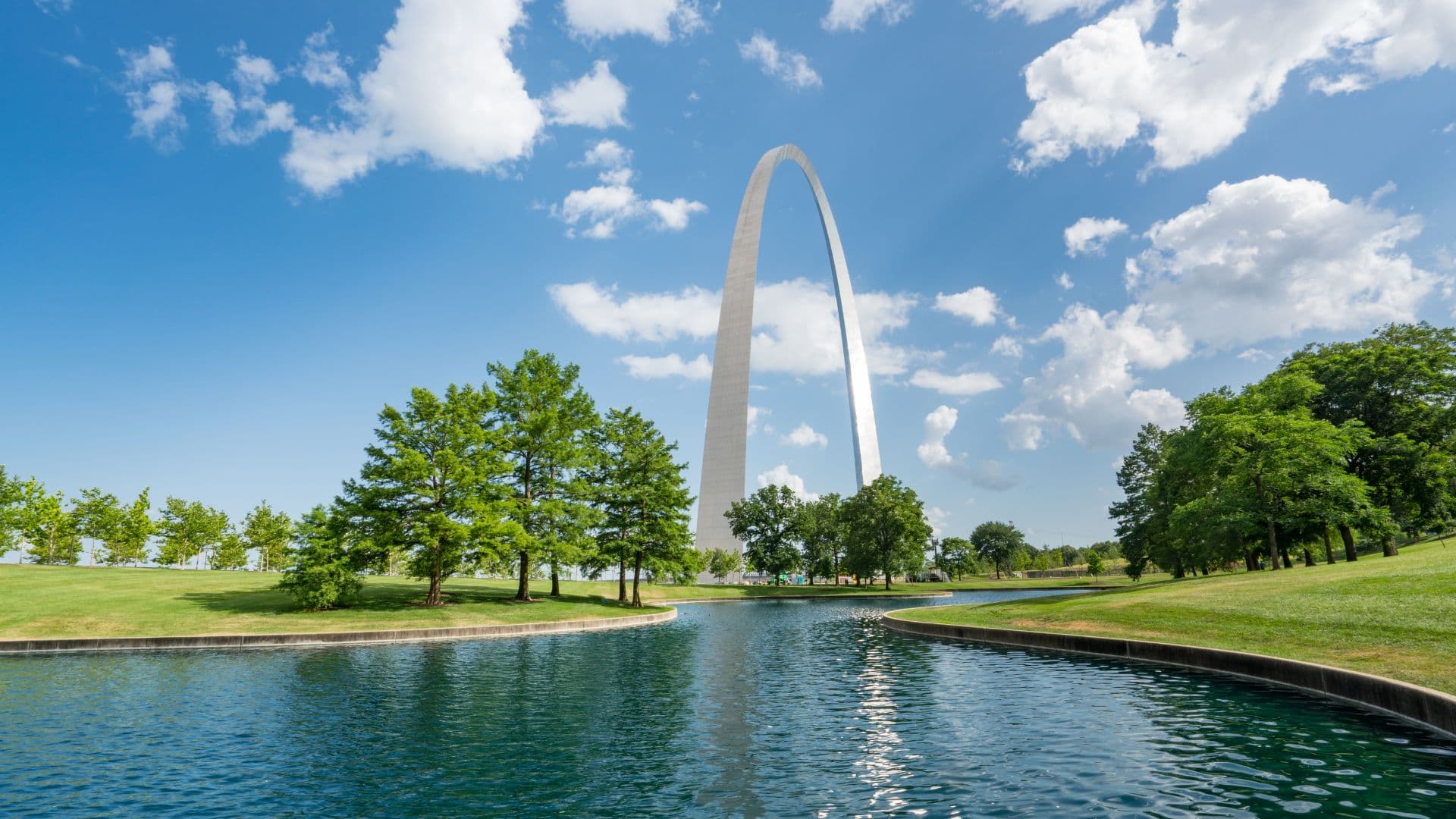 The Gateway Arch rises majestically above a serene pond and lush greenery under a blue sky with fluffy clouds.