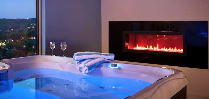 A luxurious hot tub with colorful lighting next to a modern fireplace and two glasses of wine.