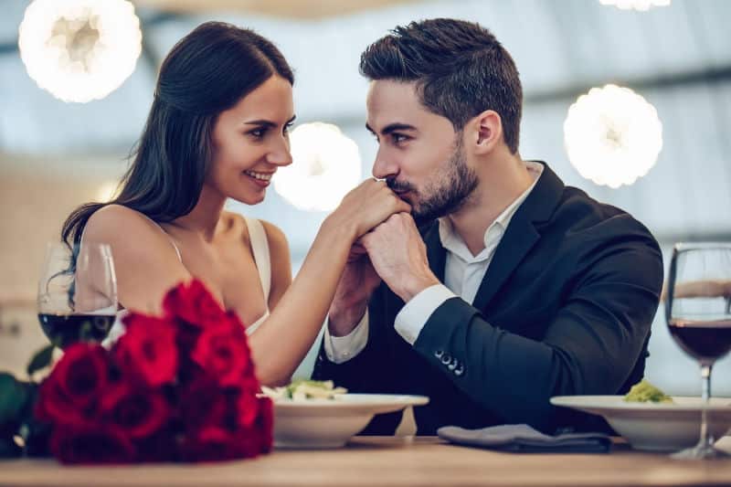 A couple gazes into each other's eyes while holding hands over a table set with food and roses.