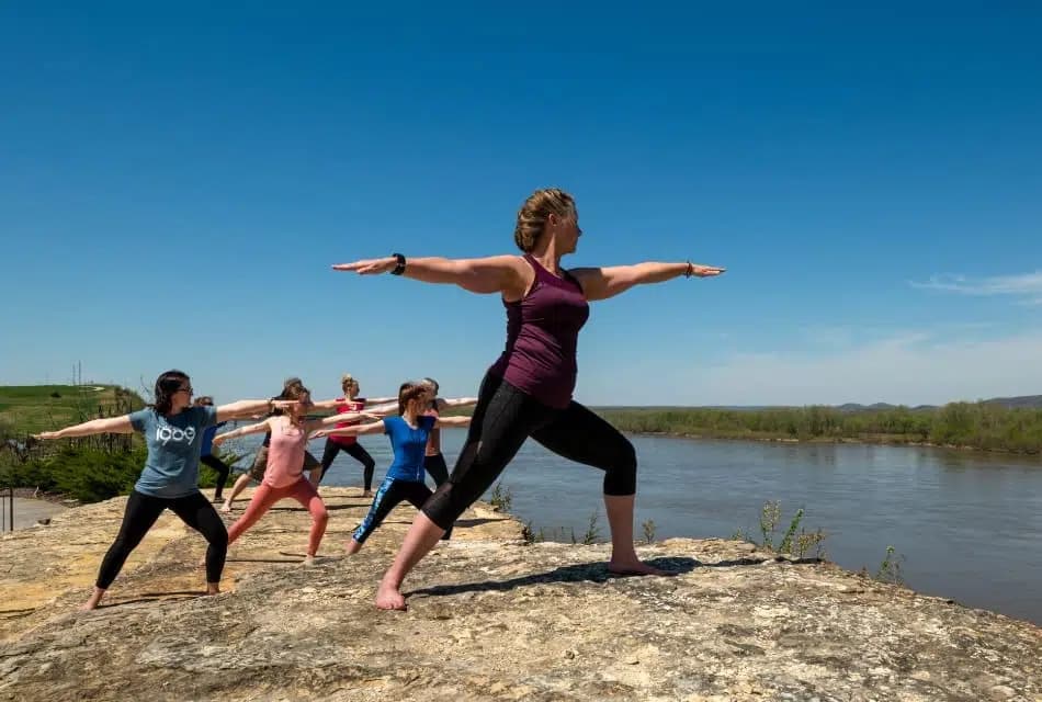 Multiple people on large rock in a yoga pose overlooking river with trees in the background