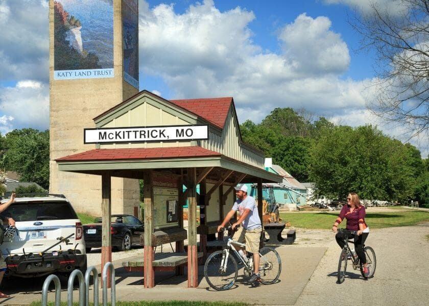 Bicyclists ride past a station sign for McKittrick, Missouri, alongside lush greenery and a prominent mural. Bicyclists ride past a station sign for McKittrick, Missouri, alongside lush greenery and a prominent mural.