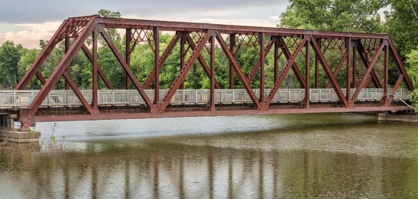 Rust-colored metal bridge over calm water surrounded by greenery.