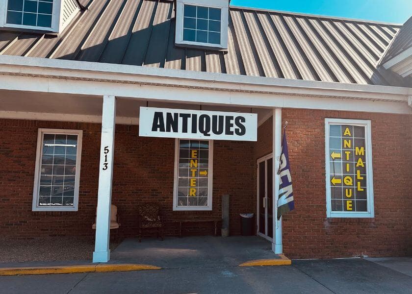 Front of an antique mall featuring a large "ANTIQUES" sign and directional arrows for entering. Front of an antique mall featuring a large "ANTIQUES" sign and directional arrows for entering.