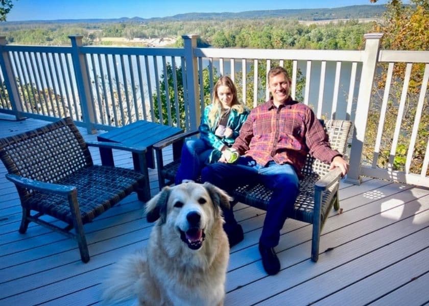 A smiling man and girl sit on a deck with a golden retriever in front of them and a scenic view in the background. A smiling man and girl sit on a deck with a golden retriever in front of them and a scenic view in the background.