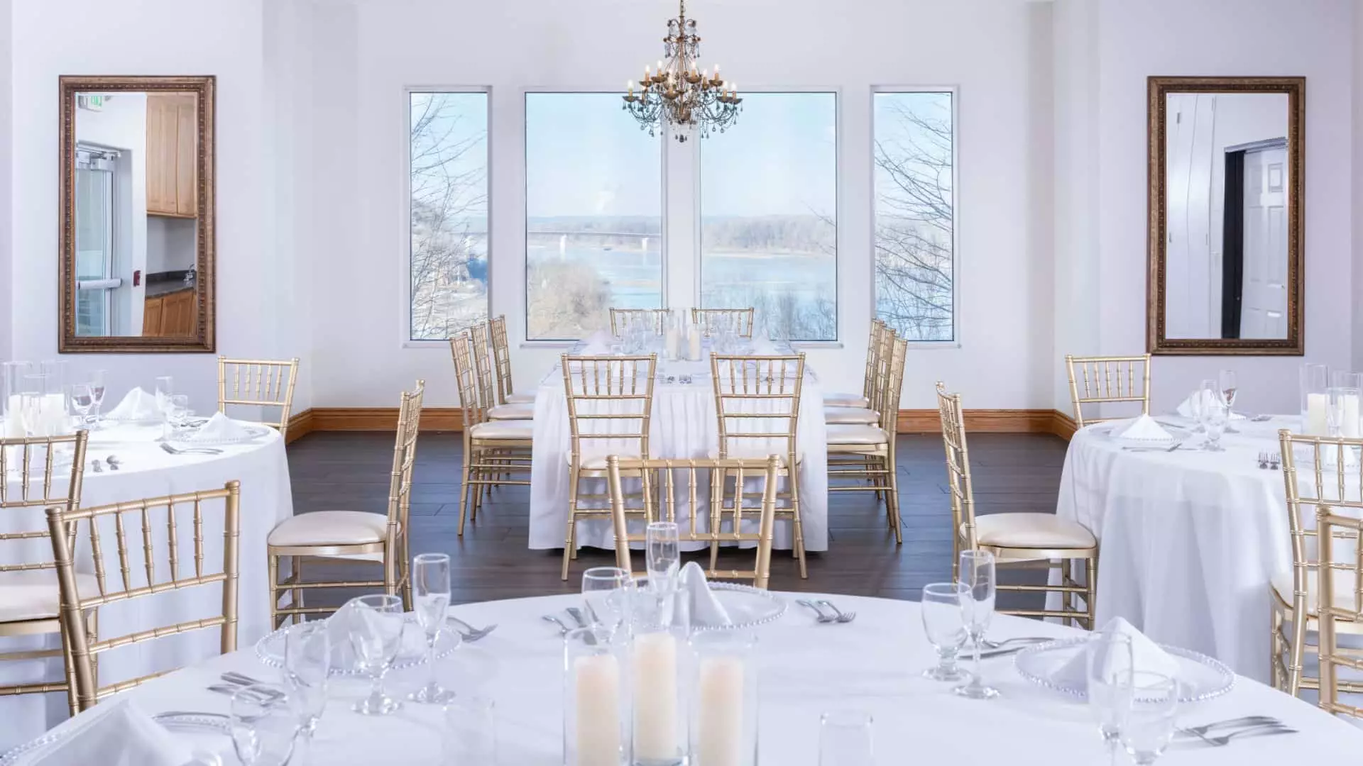 Large banquet hall with gold chairs, tables with white tablecloths, full placesettings, and windows showing the river