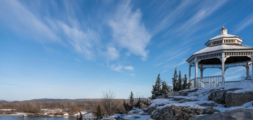A white gazebo on rocky terrain overlooking a snowy landscape and blue sky.