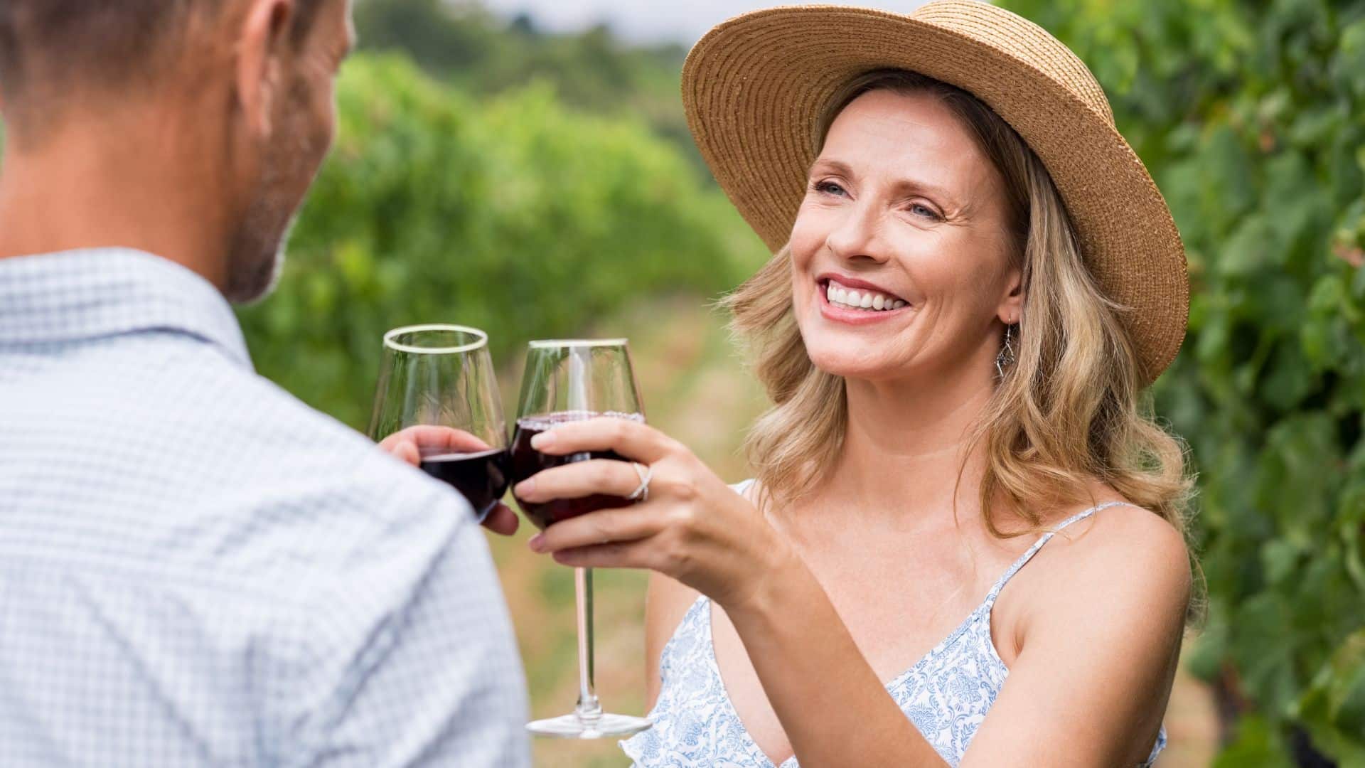 A smiling woman in a straw hat holds two glasses of red wine in a vineyard.