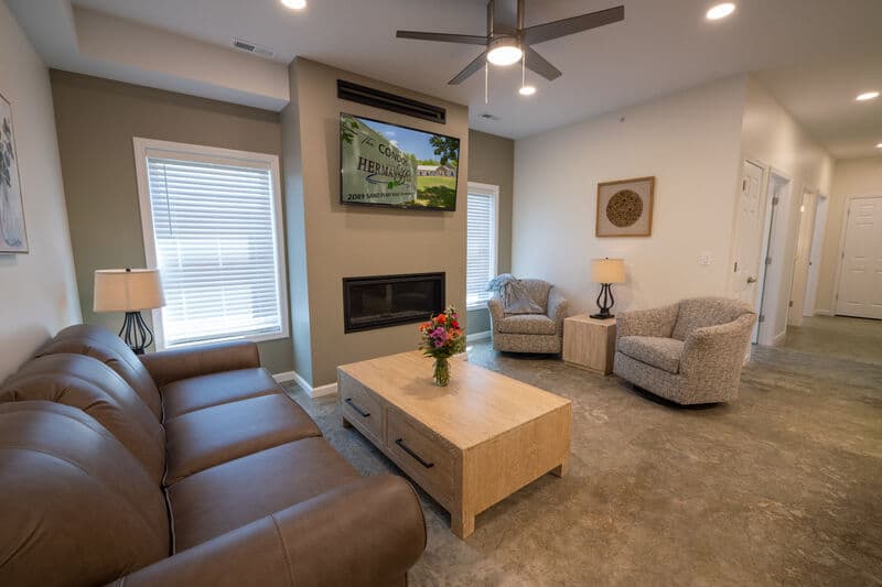 A modern living room featuring a brown leather sofa, two patterned armchairs, a wooden coffee table with a vase of flowers, and a wall-mounted TV.