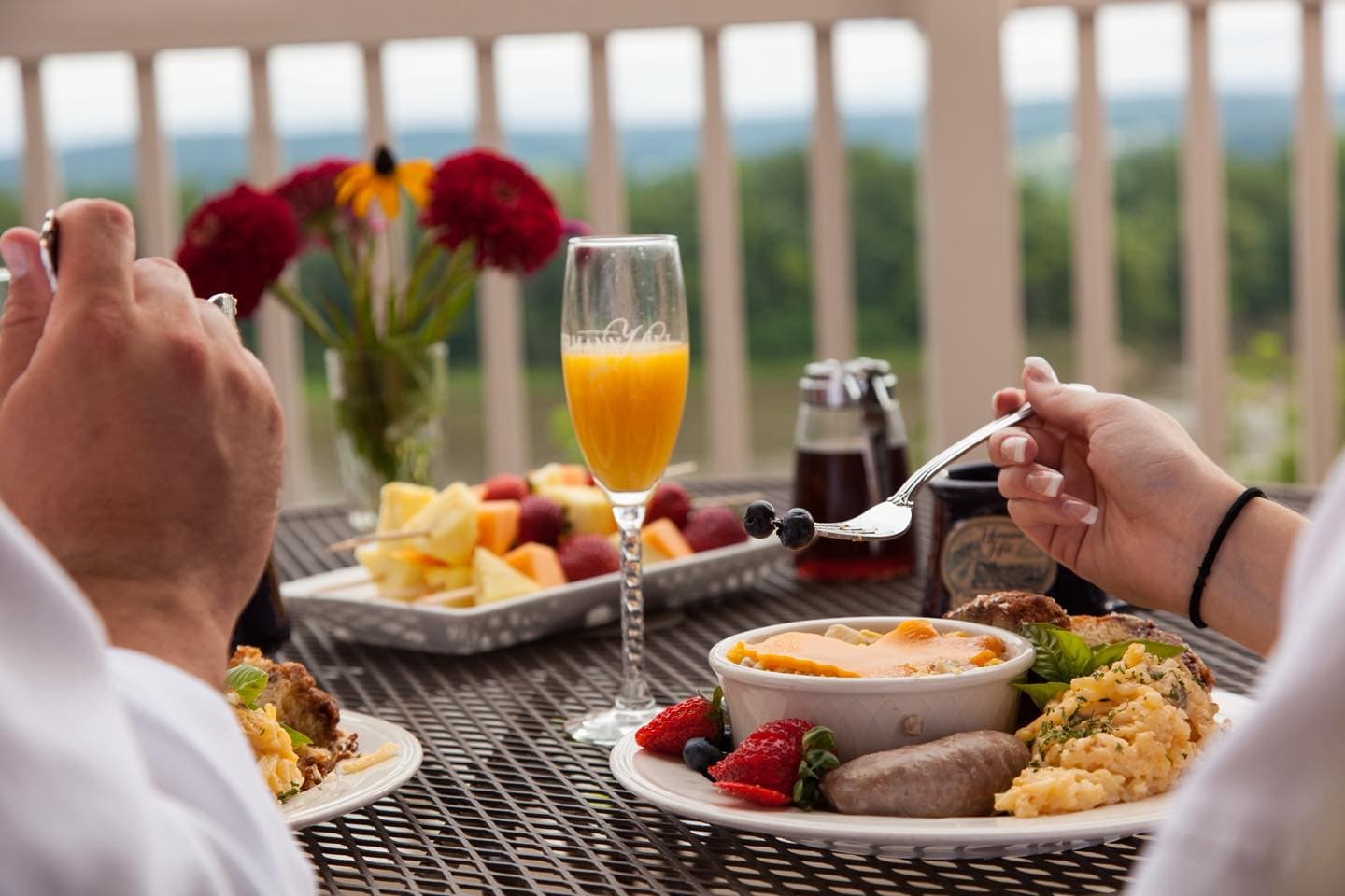 A close-up of a brunch table with two plates of food, a glass of orange juice, and fresh fruit, set against a scenic outdoor backdrop.