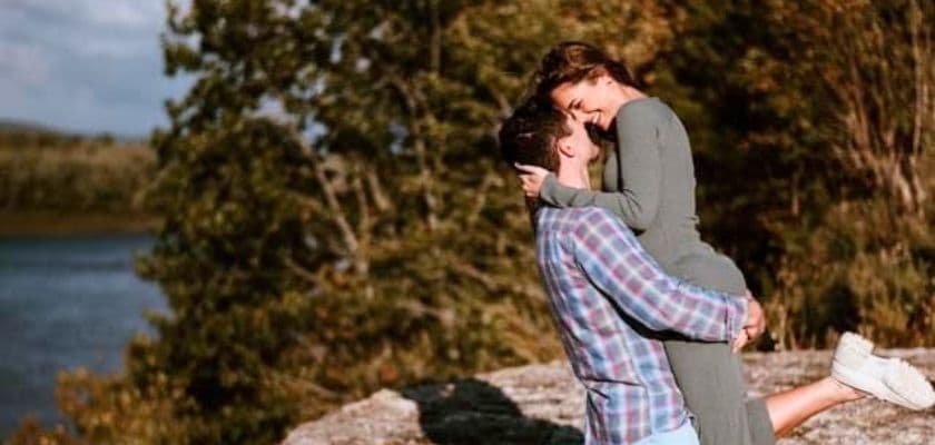 A couple embraces joyfully outdoors, with the woman lifted in the man's arms near a river.