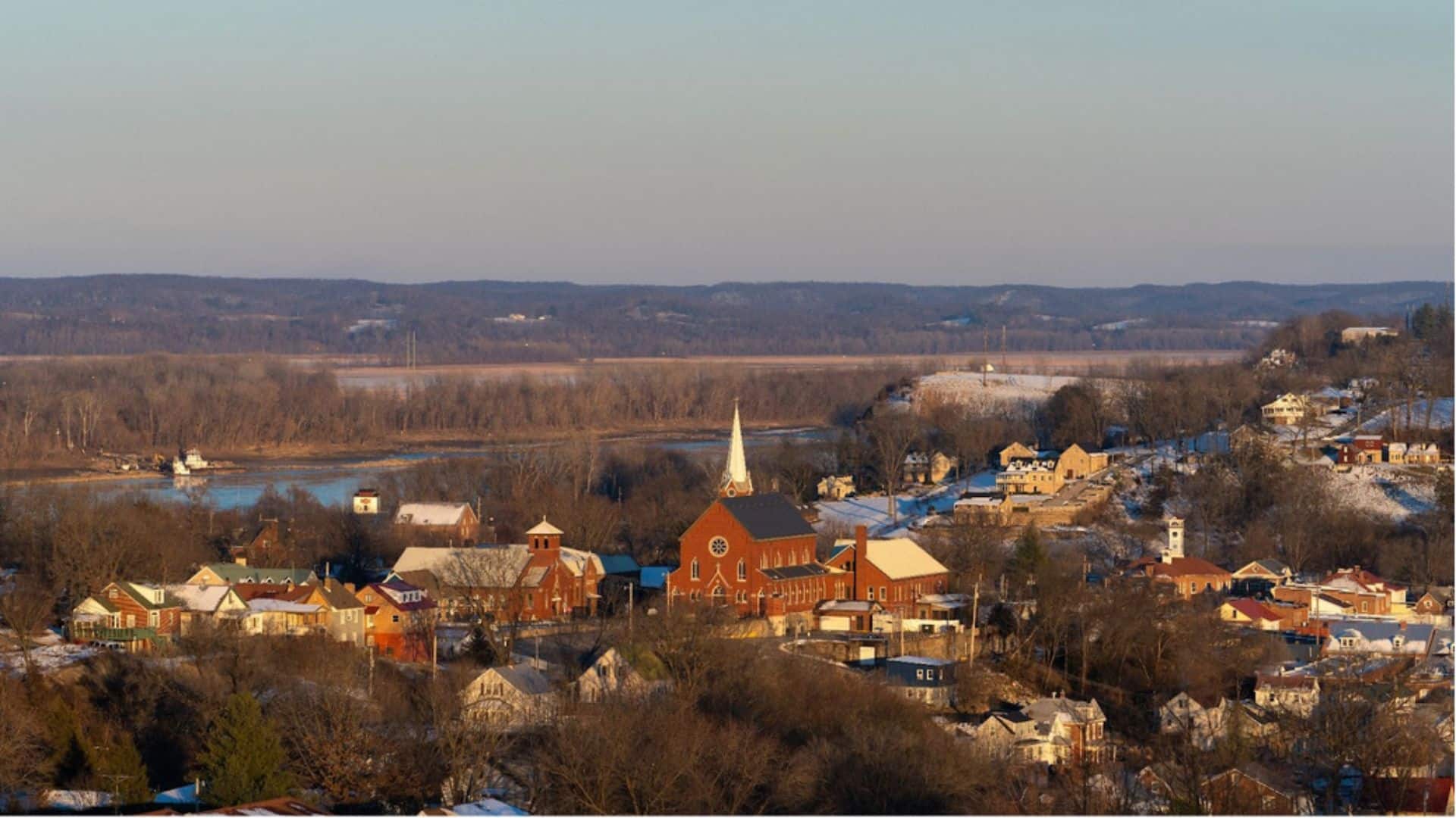 A panoramic view of a small town with a prominent church steeple, surrounded by snowy landscape and a river in the background.
