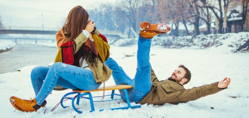 A woman on a sled laughs while a man playfully falls back in the snow beside her. A woman on a sled laughs while a man playfully falls back in the snow beside her.