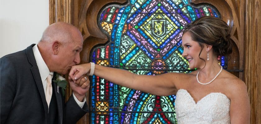 A bride and her father share a tender moment in front of a colorful stained glass window.