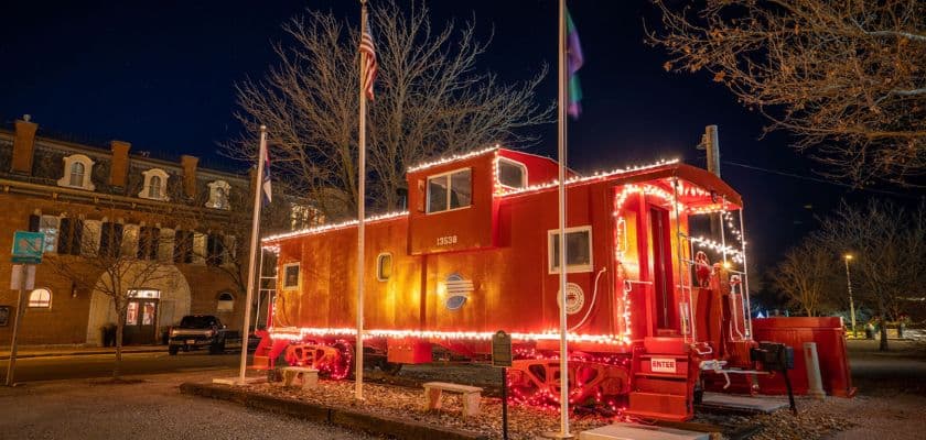 A brightly lit red caboose stands illuminated at night, surrounded by flags and trees.