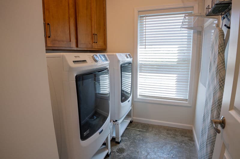 A laundry room featuring two white washing machines next to a window with blinds.