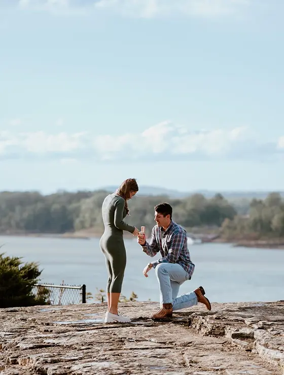 A man kneels on a rock by a lake, proposing to a woman who looks surprised and happy.