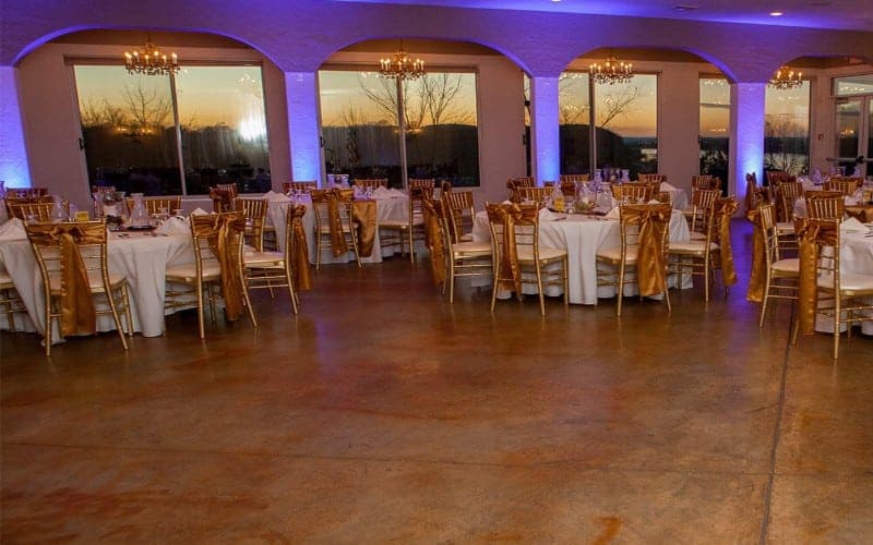 Elegant event space with round tables set for dining, accented by golden chair sashes and sunset views through large windows.