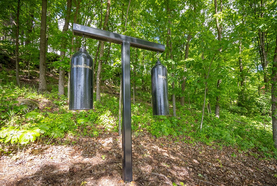 Two black gongs hanging from a black T shaped metal frame. There is a hill in the background with a forest of trees.