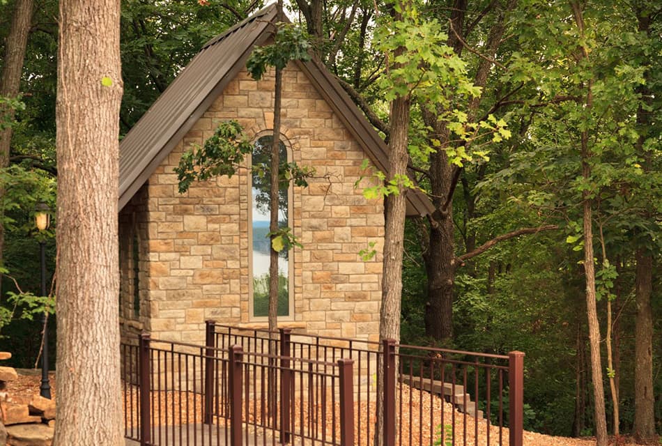 Small stone building with arched window in the woods  and lots of trees in background. Metal railed footbridge and tree trunks in foreground.