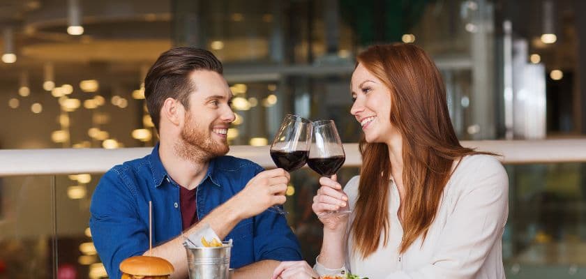 A happy couple is seated at a casual dining restaurant, toasting wine and sharing a burger, French fries, and a salad.