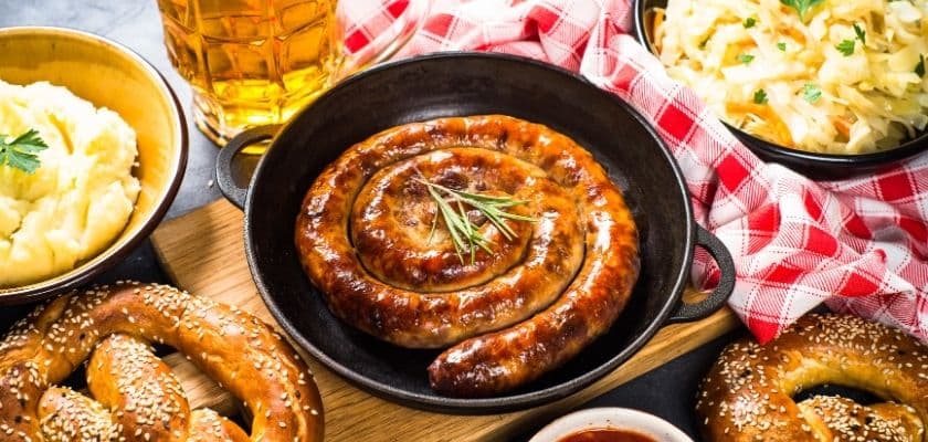 traditional german food plated on a table including sausage, beer, mashed potatoes, and a large bretzel