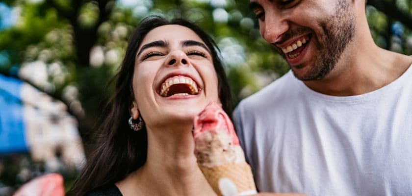 A laughing couple sharing an ice cream cone outside.