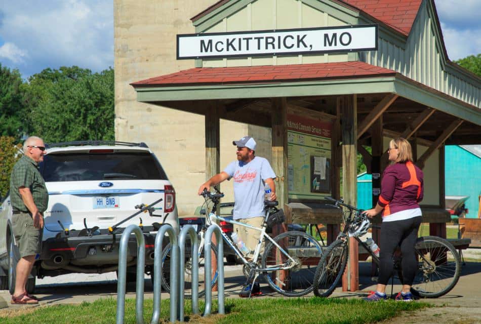 Man standing by car and two other people standing with bicycles next to shelter providing trail maps