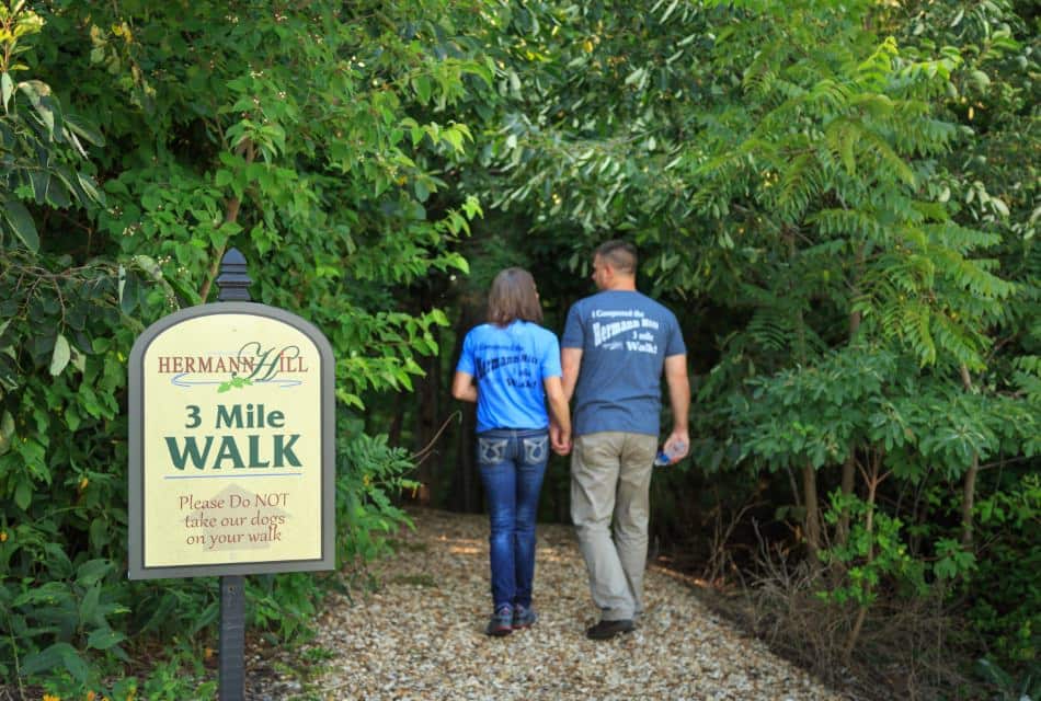 Man and woman on gravel path walking towards a trail covered with green trees