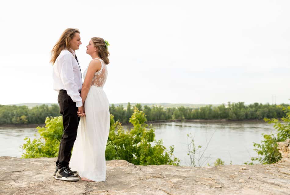 Man with white shirt and black tie holding the hands of woman in white dress with river in the background