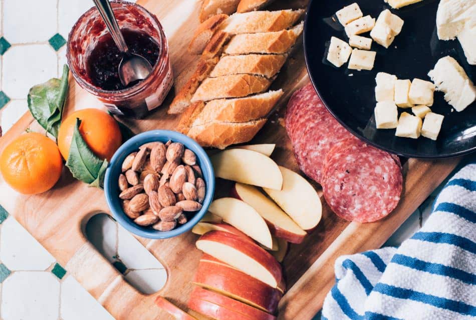 Close up view of a wooden board with sliced apples, bread, and sausage, bowl of almonds, and plate of cheese