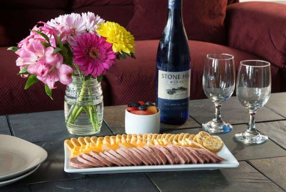 White rectangular dish with cheese, crackers, and sausage, small fruit cup, vase with pink and yellow flowers, and bottle of wine