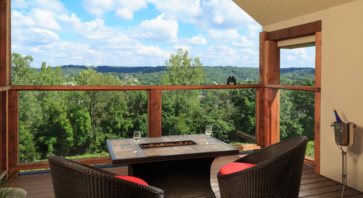 Enclosed deck with dark wicker patio furniture, red cushions, table fire pit, and view of green trees and hills in the background