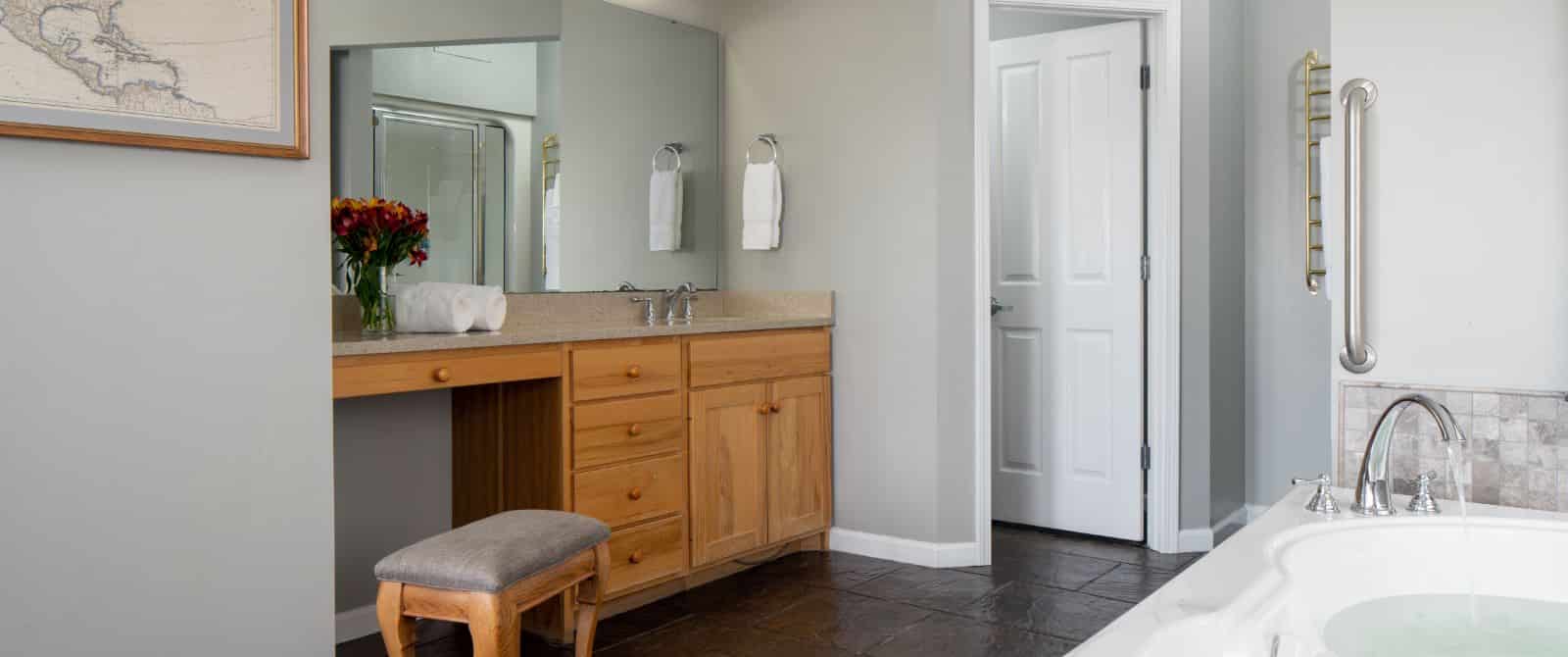Large bathroom with light gray walls, tiled flooring, wooden vanity with quartz top, and jetted tub