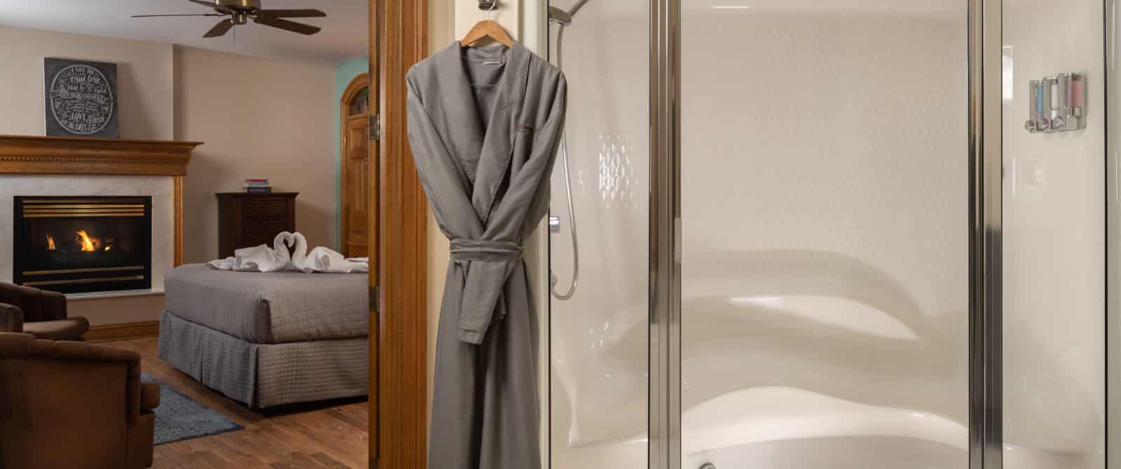 Large walkin shower, gray robe hanging nearby, and view into bedroom with cream walls, wooden flooring, fireplace, and gray bedding