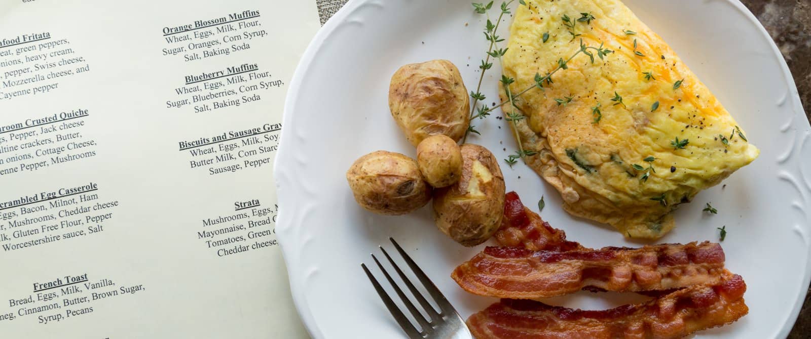 Close up view of breakfast menu and white plate with omelette, bacon, and potatoes