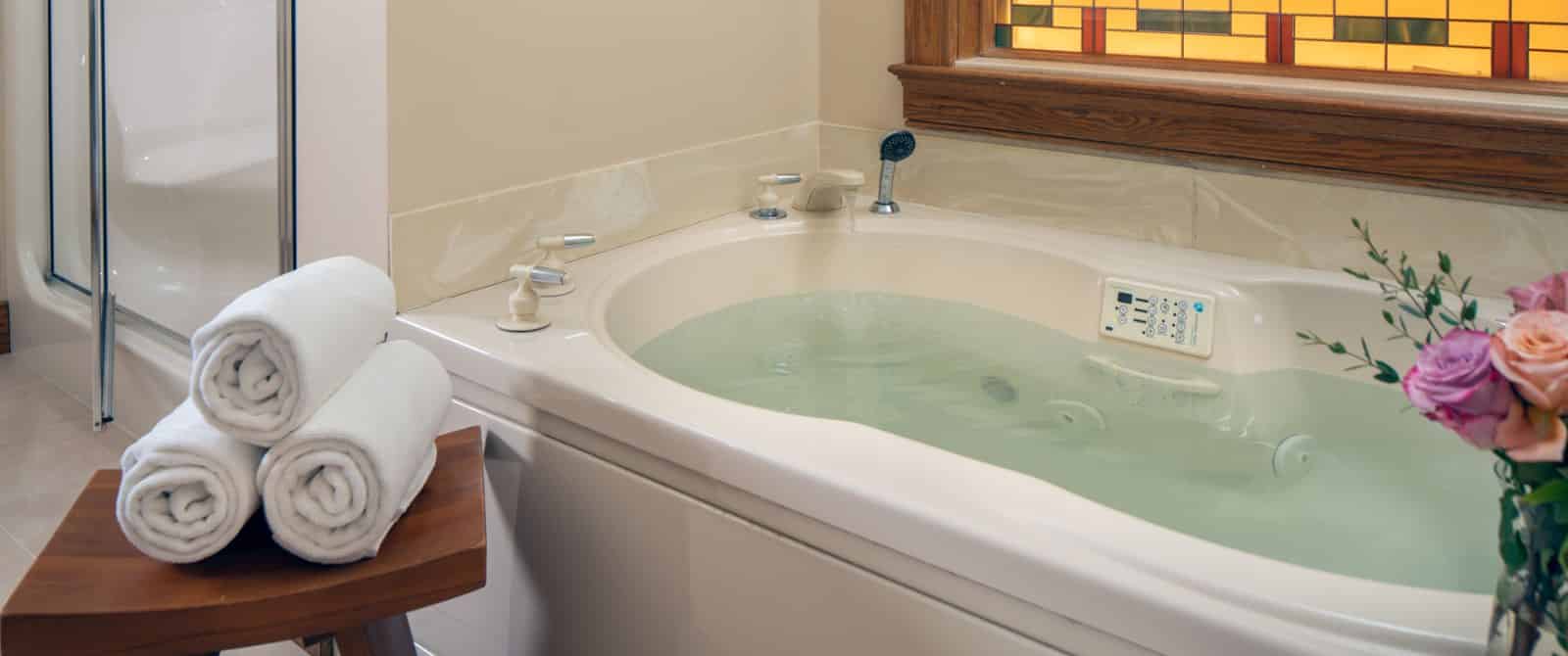 Close up view of large jetted tub filling with water and a small wooden stool with three white rolled towels