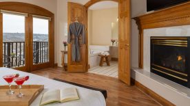 Large bedroom suite with cream walls, wooden flooring, fireplace, white bedding, and view into bathroom