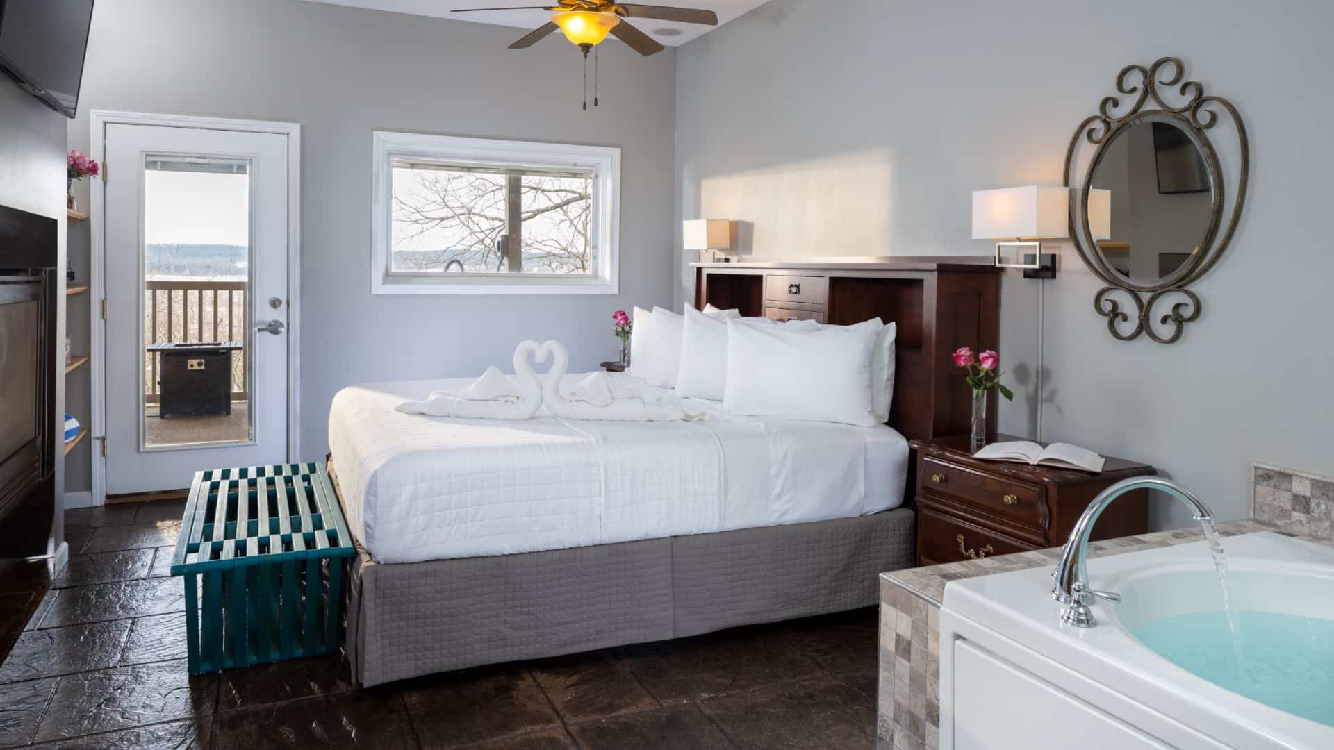 Large bedroom suite with light gray walls, tiled flooring, wooden headboard, white bedding, fireplace, and jetted tub