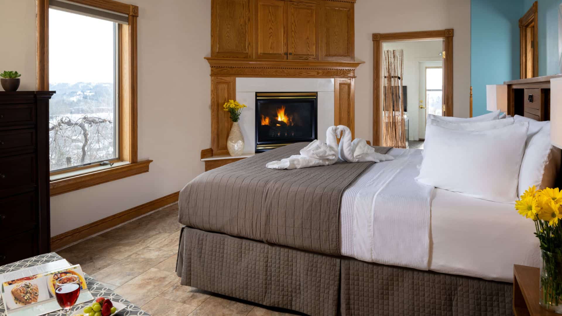 Large bedroom suite with cream and turquoise walls, tiled flooring, white bedding, gray comforter, large fireplace, and view into bathroom