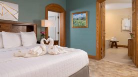 Large bedroom suite with teal walls, tiled flooring, wooden headboard, white bedding, and view into bathroom