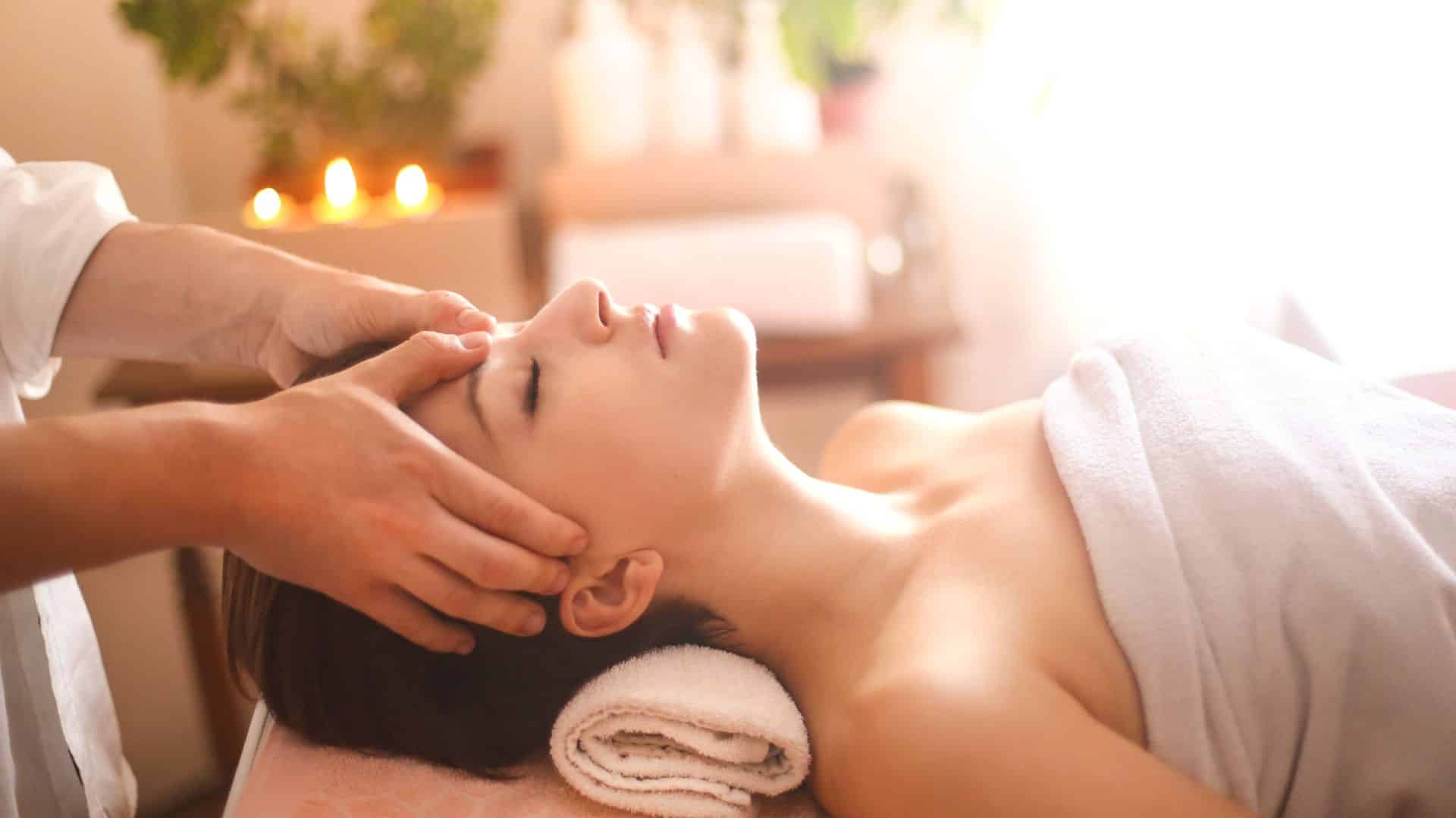 woman with brown hair wearing a white towel receiving a forehead massage with plants, lotions, and candles in the background