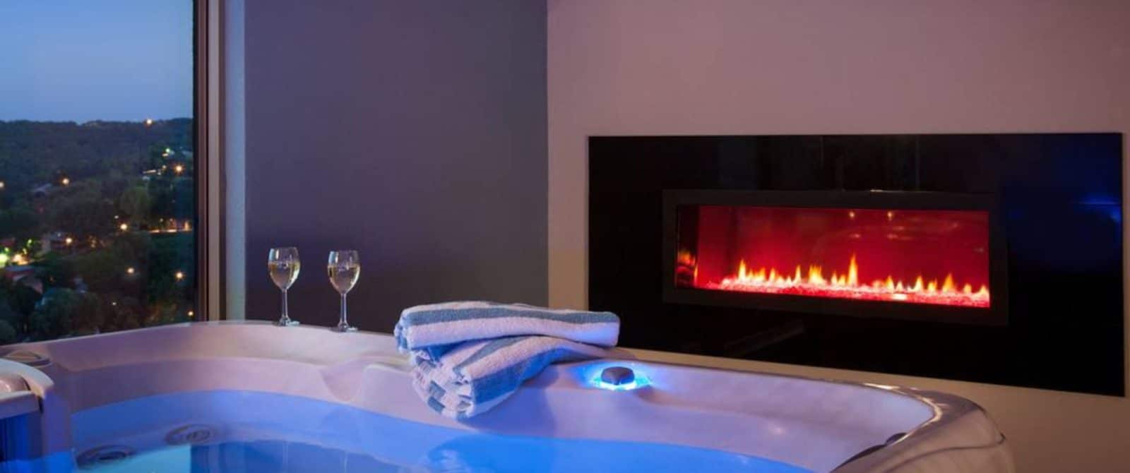 Jetted tub in front of a linear gas fireplace and a window overlooking the town of Hermann at night.